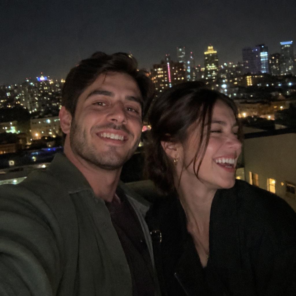 A candid selfie-style photo of a couple on a rooftop at night, city lights behind them