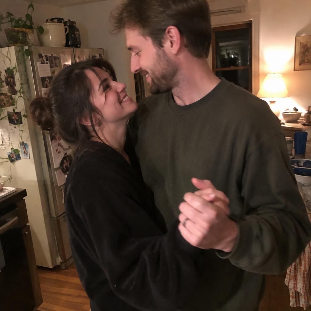 A candid snapshot of a couple slow dancing in their kitchen at night under warm lamplight